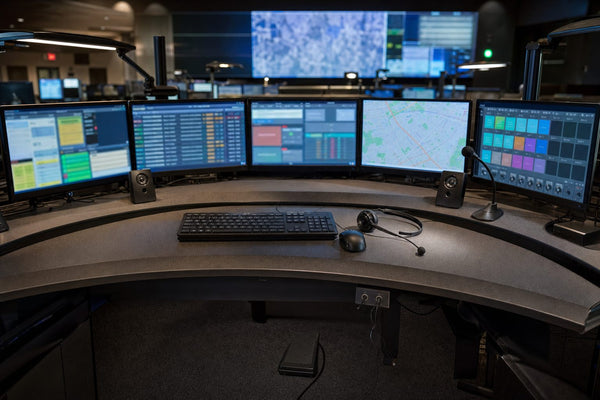 A 911 dispatch position with CAD and map monitors, a radio console screen, desk speakers, a gooseneck microphone, a headset on the desk and a foot pedal underneath